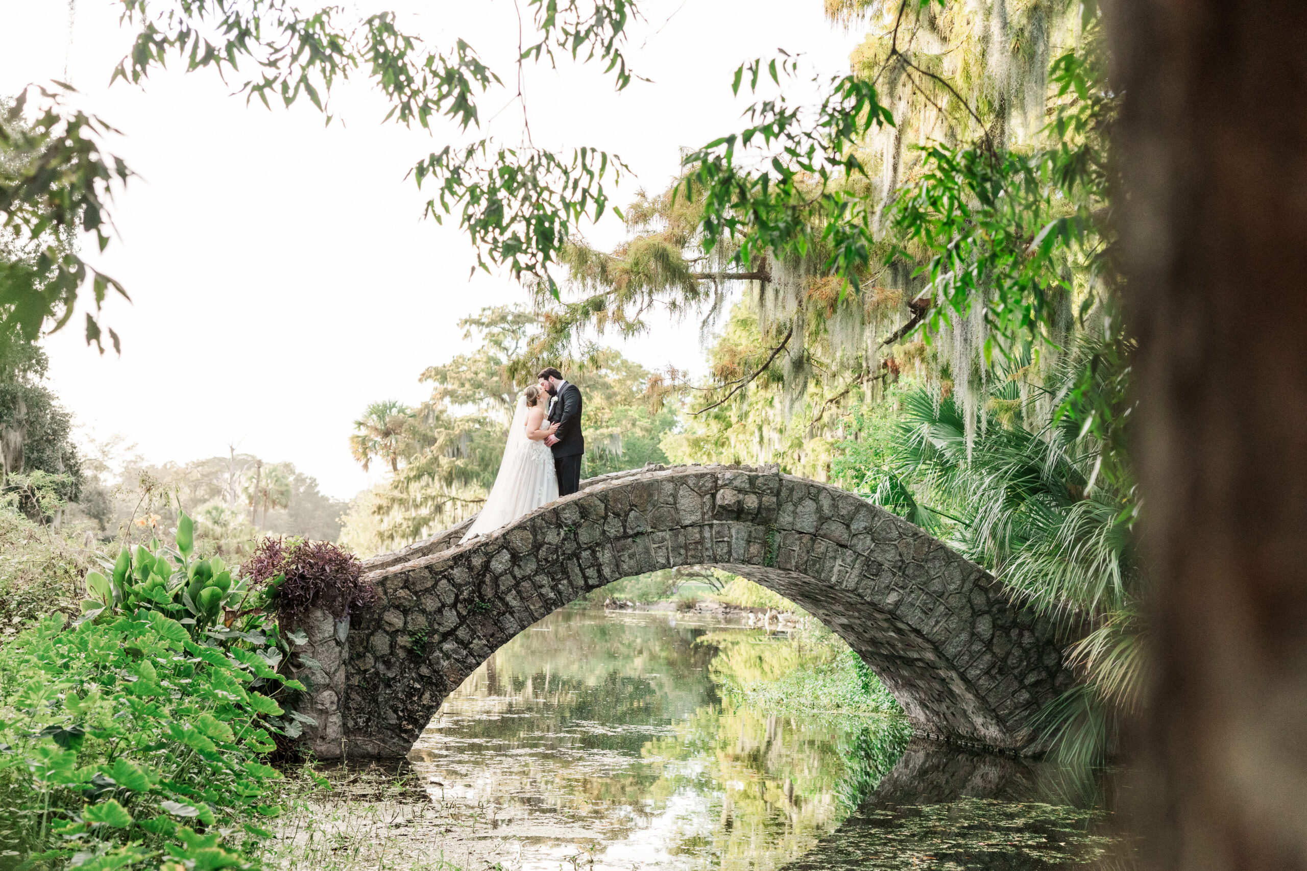 A bride and groom share an intimate moment on a historic stone bridge surrounded by lush greenery and Spanish moss in City Park Louisiana. The bride wears a flowing white gown, and the groom is dressed in a classic black suit as they embrace in this romantic outdoor wedding setting.