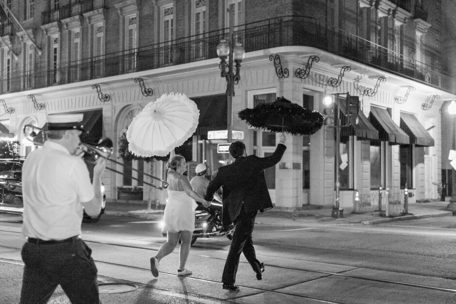 A newlywed couple joyfully parades through the streets of New Orleans at night, holding traditional second line umbrellas while a trombone player leads the way. The groom wears a classic black suit, and the bride stuns in a short white dress as they celebrate their wedding in true New Orleans style.