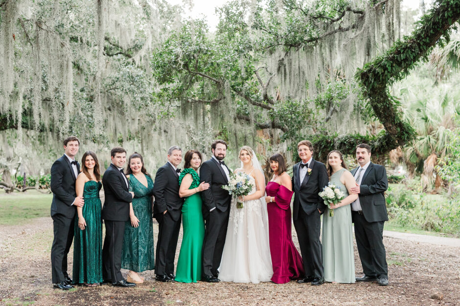 A joyful wedding portrait of the bride and groom surrounded by their family and wedding party, dressed in elegant formal attire, under a canopy of Spanish moss-covered trees in City Park Loussiana. The lush greenery and natural backdrop create a timeless and romantic setting.