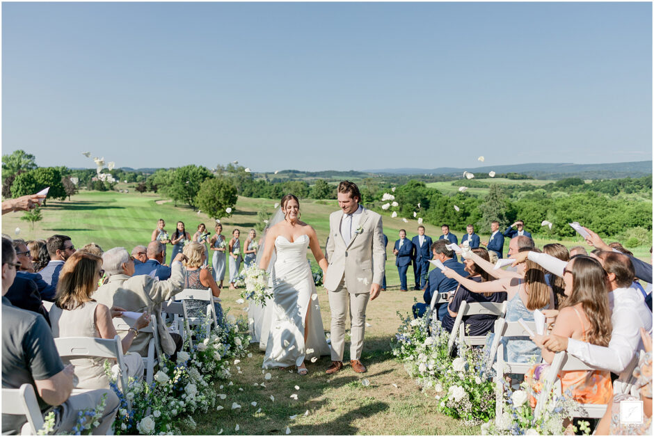 Bride in a white gown and groom in a tan suit walk down an outdoor aisle, smiling as guests toss white petals in celebration against a scenic countryside backdrop with their marriage license.